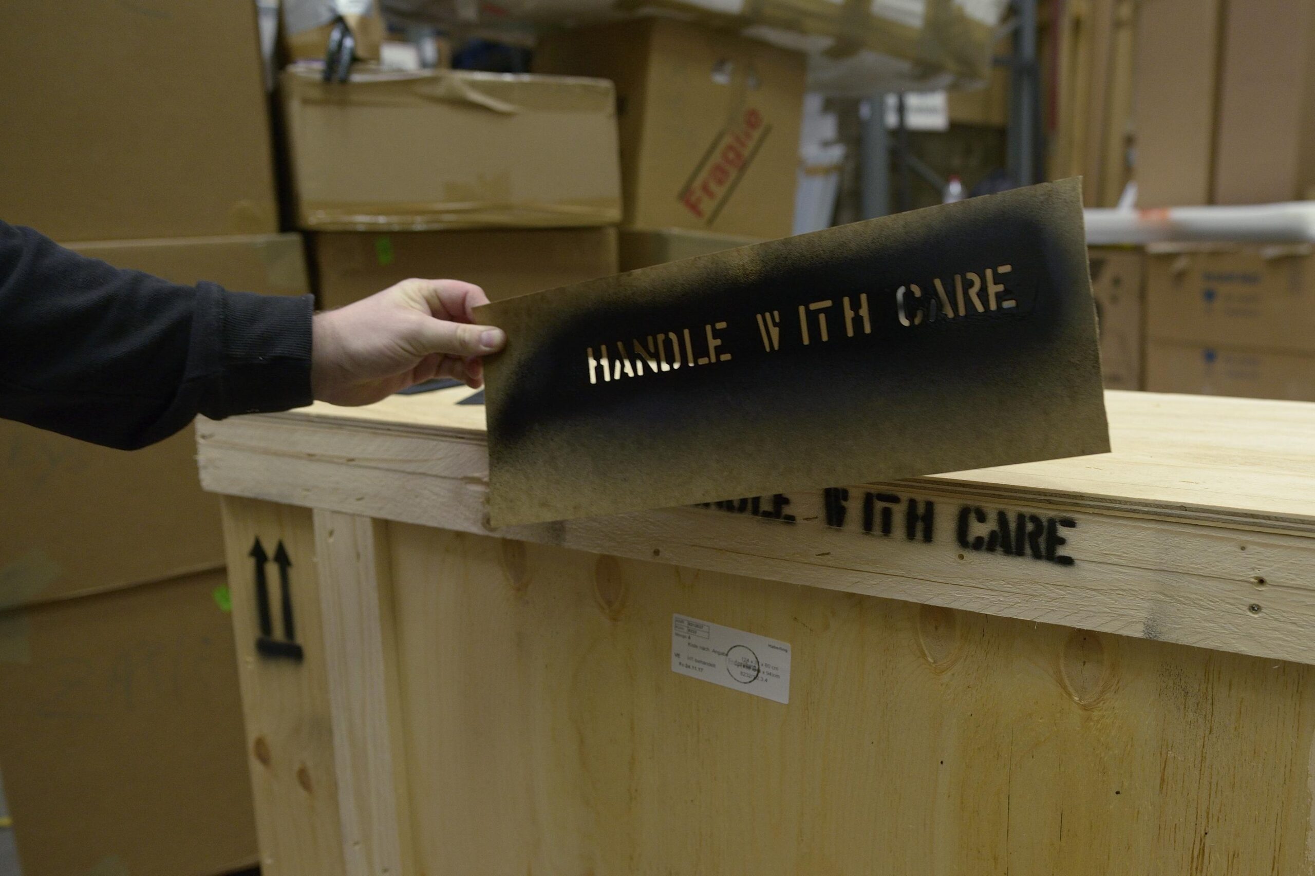 Eine Hand hält eine Schablone mit der Aufschrift „Handle with care“ über einer Holzkiste. Die Worte sind auf die Kiste gesprüht. Im Hintergrund sind Kartons zu sehen.
