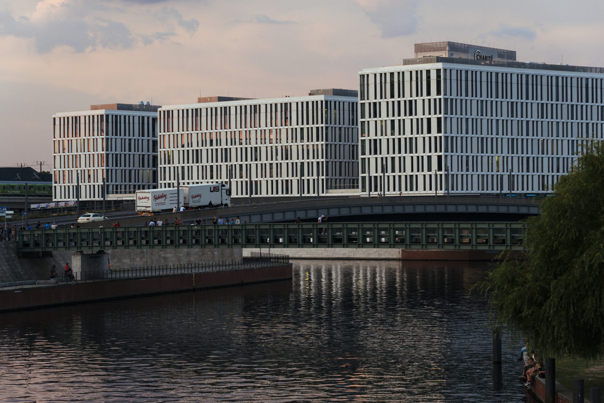 Moderne weiße Gebäude mit vertikalen Fenstern ragen über einer belebten Brücke über dem Wasser und spiegeln den ruhigen Himmel am frühen Abend wider. Menschen und Haberling Lkw bewegen sich darüber hinweg.