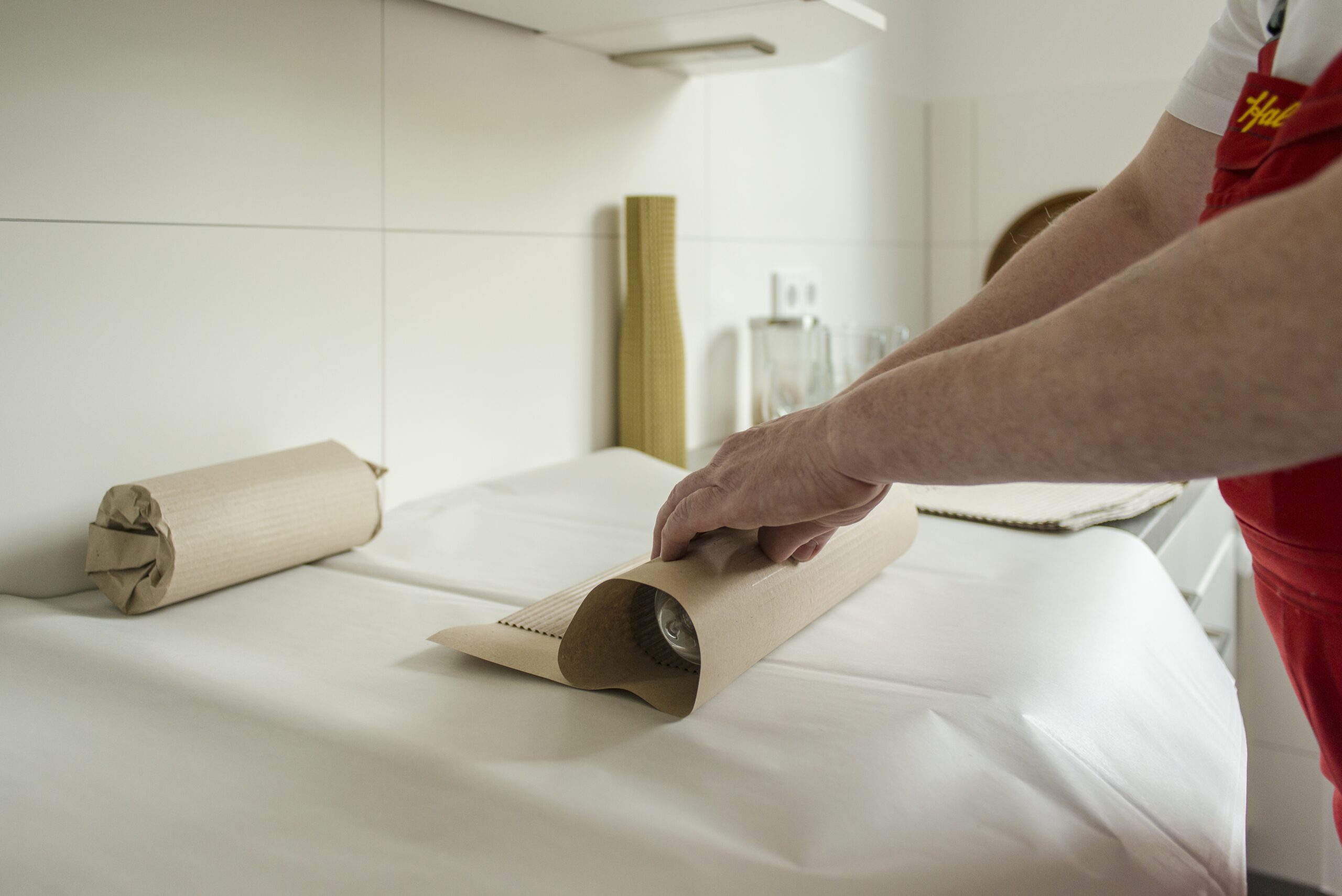 Umzugshelfer in einer roten Uniform wickelt eine Glasflasche sorgfältig in braunes Papier auf einer weißen Fläche in einer Küche ein.