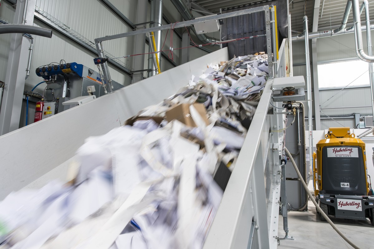 Industrielle Umgebung mit einem Förderband, das zerkleinertes Papier in einer Recyclinganlage transportiert.