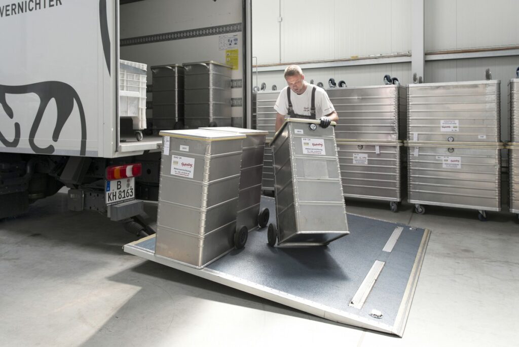 Ein Mitarbeiter entlädt Metallcontainer mithilfe einer an einem Lkw befestigten Rampe in einem Lagerhaus.