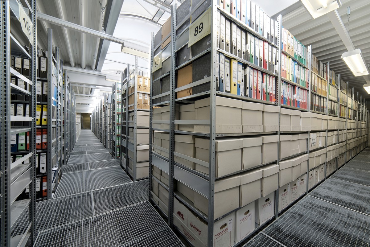 Reihen von Metallregalen, gefüllt mit ordentlich sortierten Ordnern und Kartons, in einem hell beleuchteten Archivraum mit hoher, gewölbter Decke.
