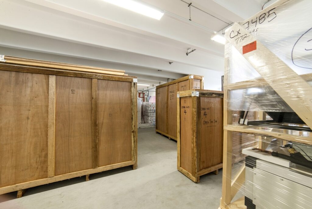 Ein Lagerbereich mit großen Holzkisten, umgeben von verpackten Gegenständen auf Paletten. Der Raum ist hell, übersichtlich und industriell gestaltet.