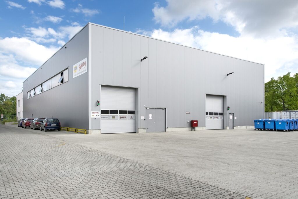 Ein modernes, graues Industriegebäude mit zwei geschlossenen Garagentoren und mehreren geparkten Autos. Auf der rechten Seite stehen blaue Recyclingbehälter.