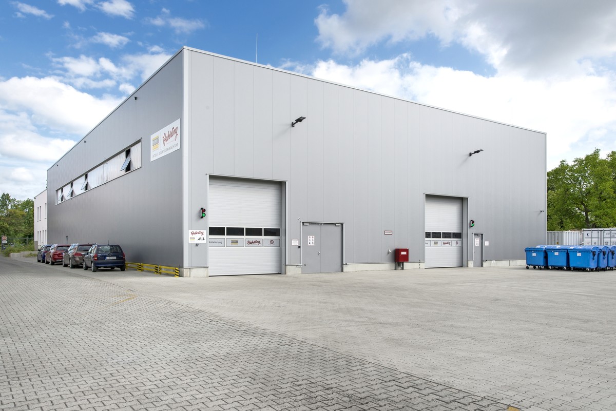 Ein modernes, graues Industriegebäude mit zwei geschlossenen Garagentoren und mehreren geparkten Autos. Auf der rechten Seite stehen blaue Recyclingbehälter.