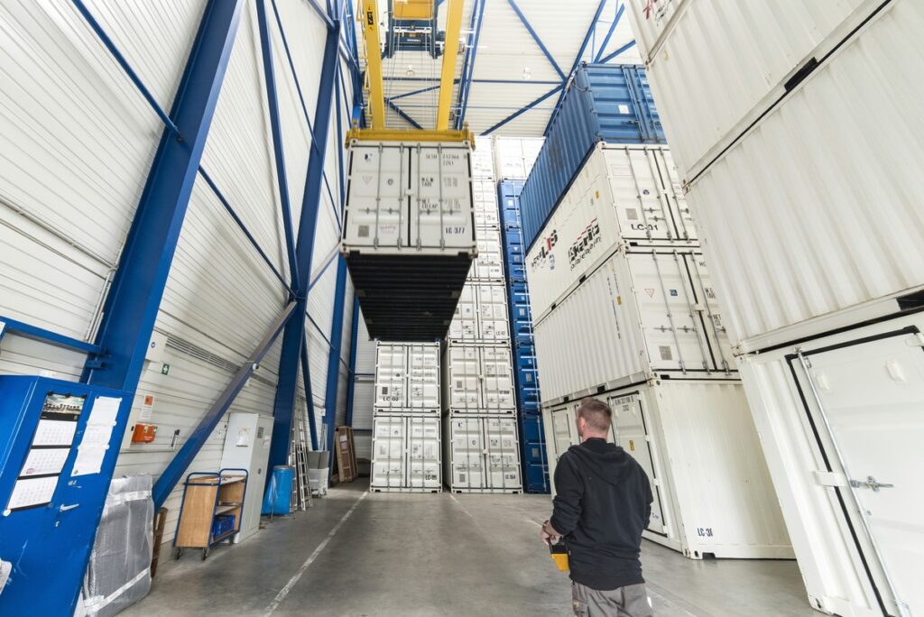 Ein Lagerinneres mit gestapelten Versandcontainern und einem Mann in einem schwarzen Kapuzenpulli, der eine Fernbedienung bedient und einen Kran überwacht, der einen Container hebt.