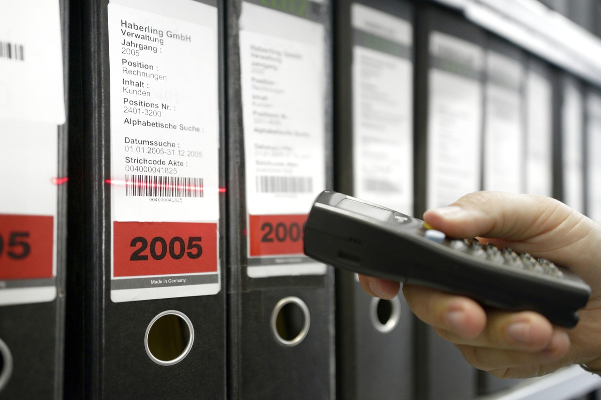 Eine Hand scannt einen Ordner mit der Aufschrift „2005” mit einem Barcode-Lesegerät. Ein roter Laser markiert den Code auf weißen Etiketten in einer Reihe ordentlicher Büroordner.