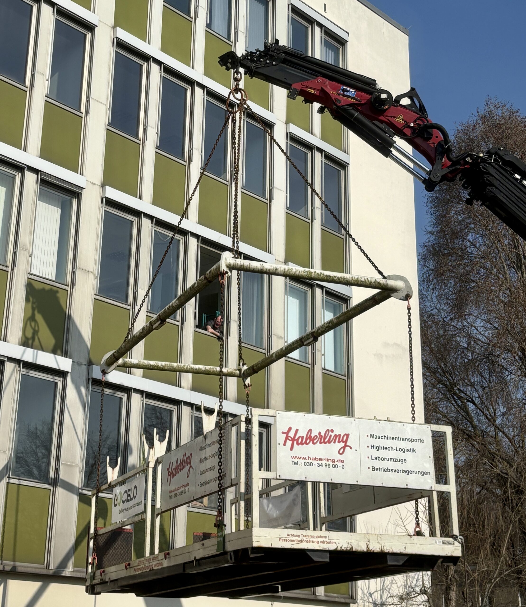 Ein Kran hebt ein großes Metallgerüst vor einem Gebäude mit grünen und weißen Paneelen an. Eine Person schaut aus einem Fenster.