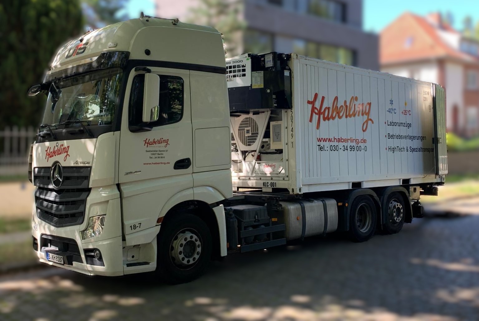 Ein weißer Lkw mit dem Schriftzug „Haberling“ steht auf einer Straße. Der Lkw transportiert einen Container. Im Hintergrund sind Bäume und Gebäude zu sehen.