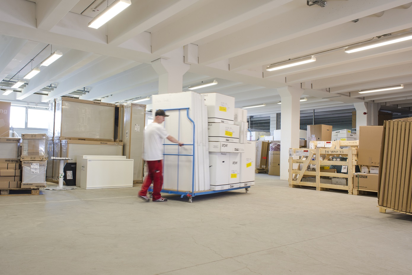 Eine Person in einem Lager schiebt einen mit großen, weißen Kartons beladenen Wagen. Um sie herum stehen Stapel von verpackten Waren.